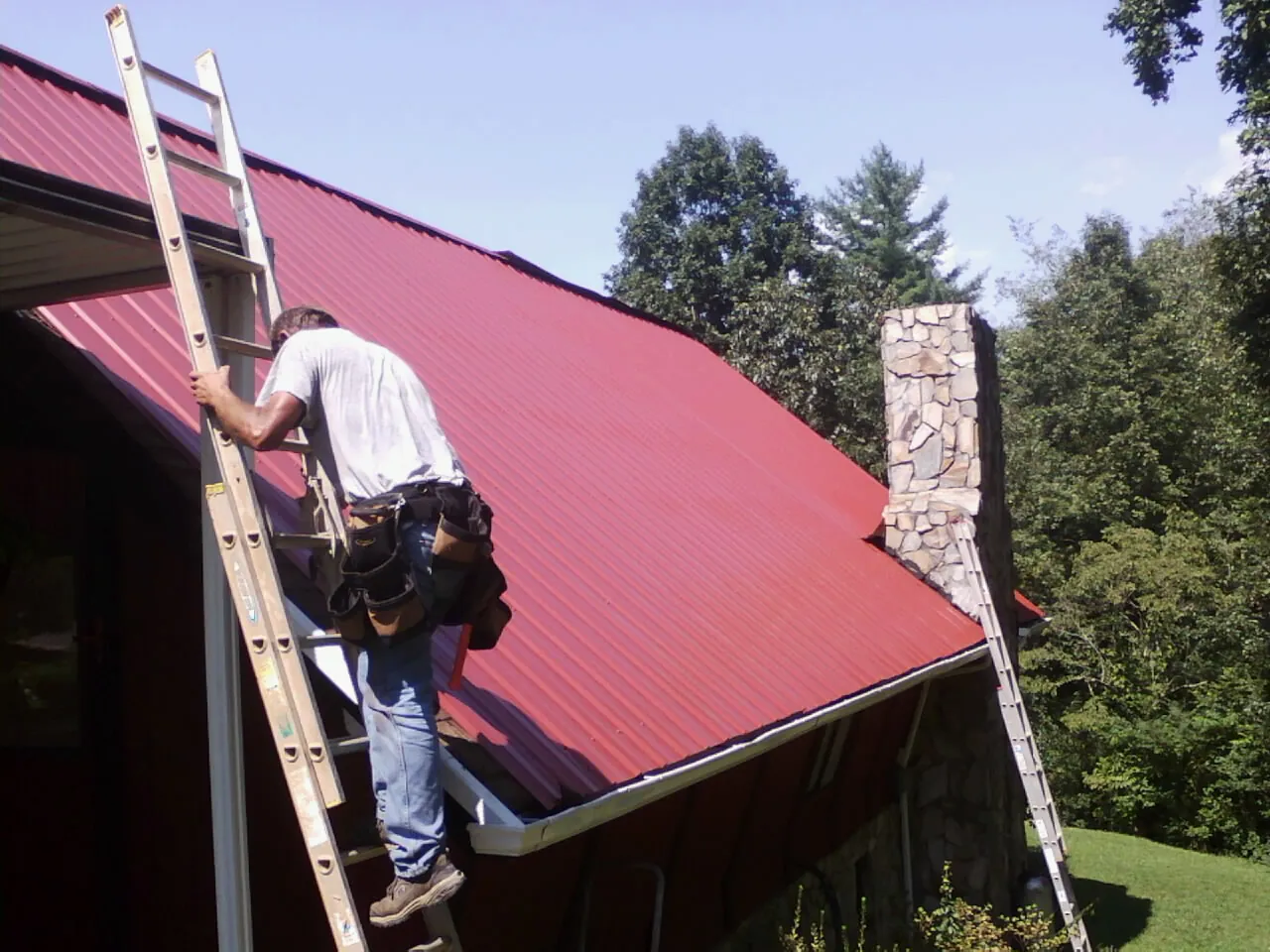 Roofer on a ladder beside a red metal roof and stone chimney — Soffit & Fascia Repair by Roofing Brothers in Rosine